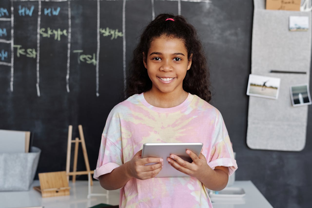 Cheerful girl holding a tablet in a modern classroom with a chalkboard background.