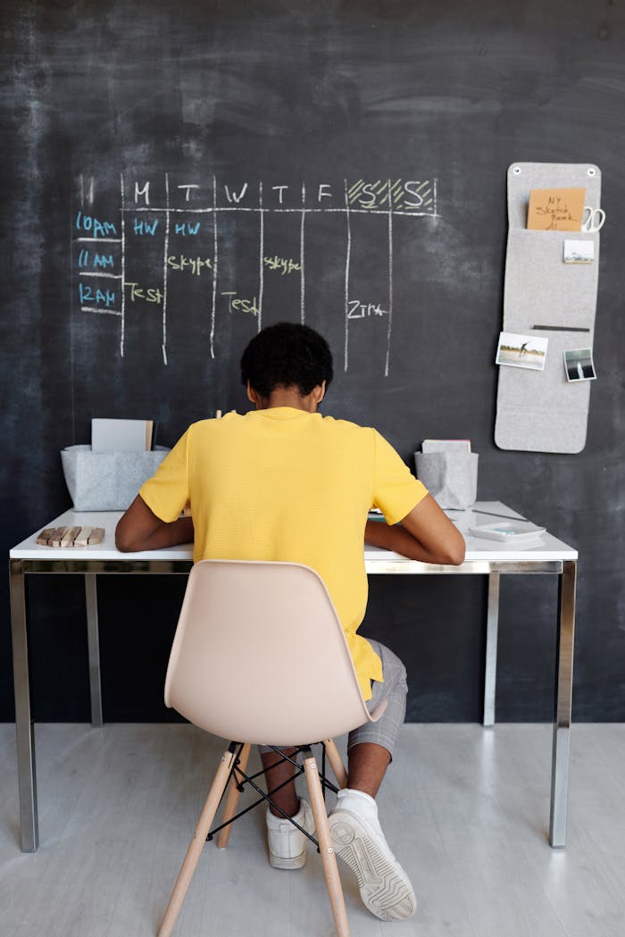 A teenager focuses on studying at a desk, with a blackboard displaying a schedule in the background.